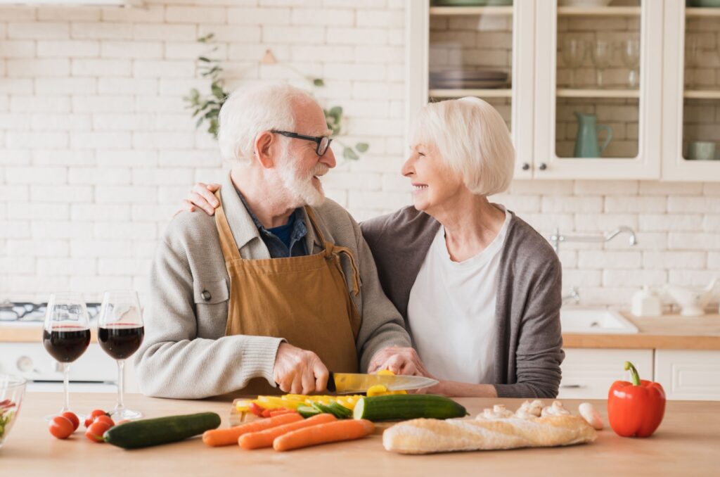 An older couple smiling at one another in their kitchen while dicing vegetables for a denture-friendly recipe.