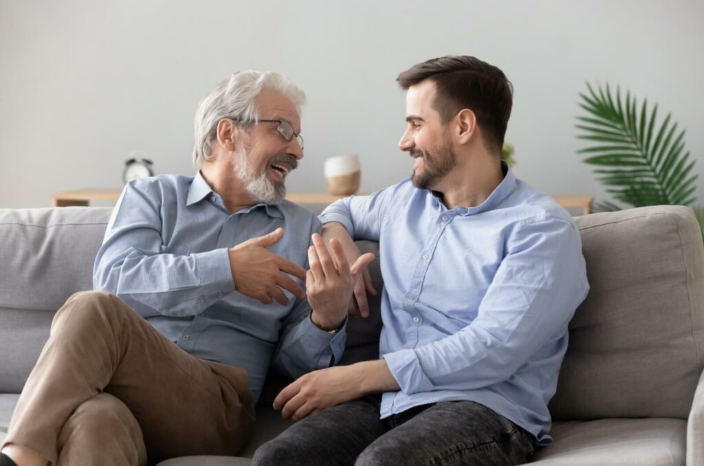 An adult child talking to their smiling older parent on the couch about the move to a retirement home.
