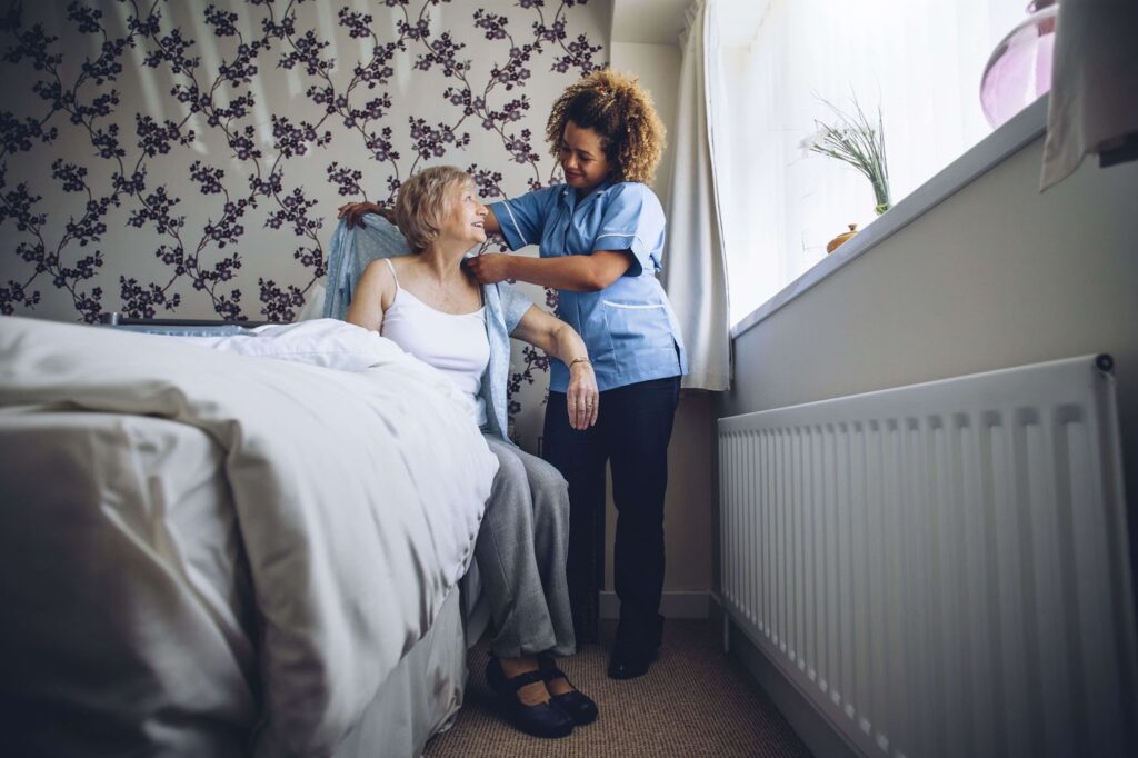 A female caregiver carefully dresses up an elderly woman seated on her bed,