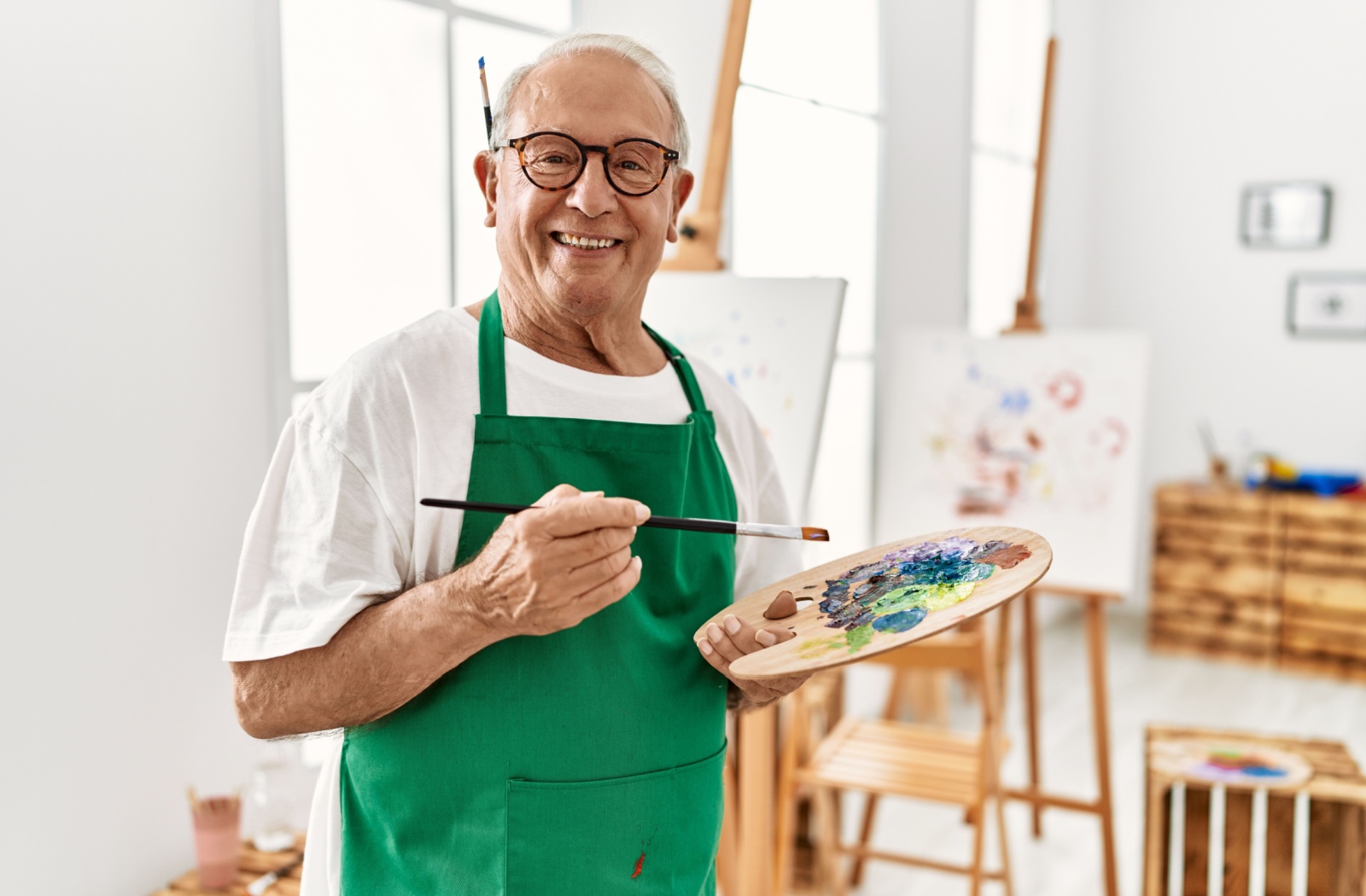 A smiling older adult in an art studio in senior living, holding up a paint palette in front of their newest painting.