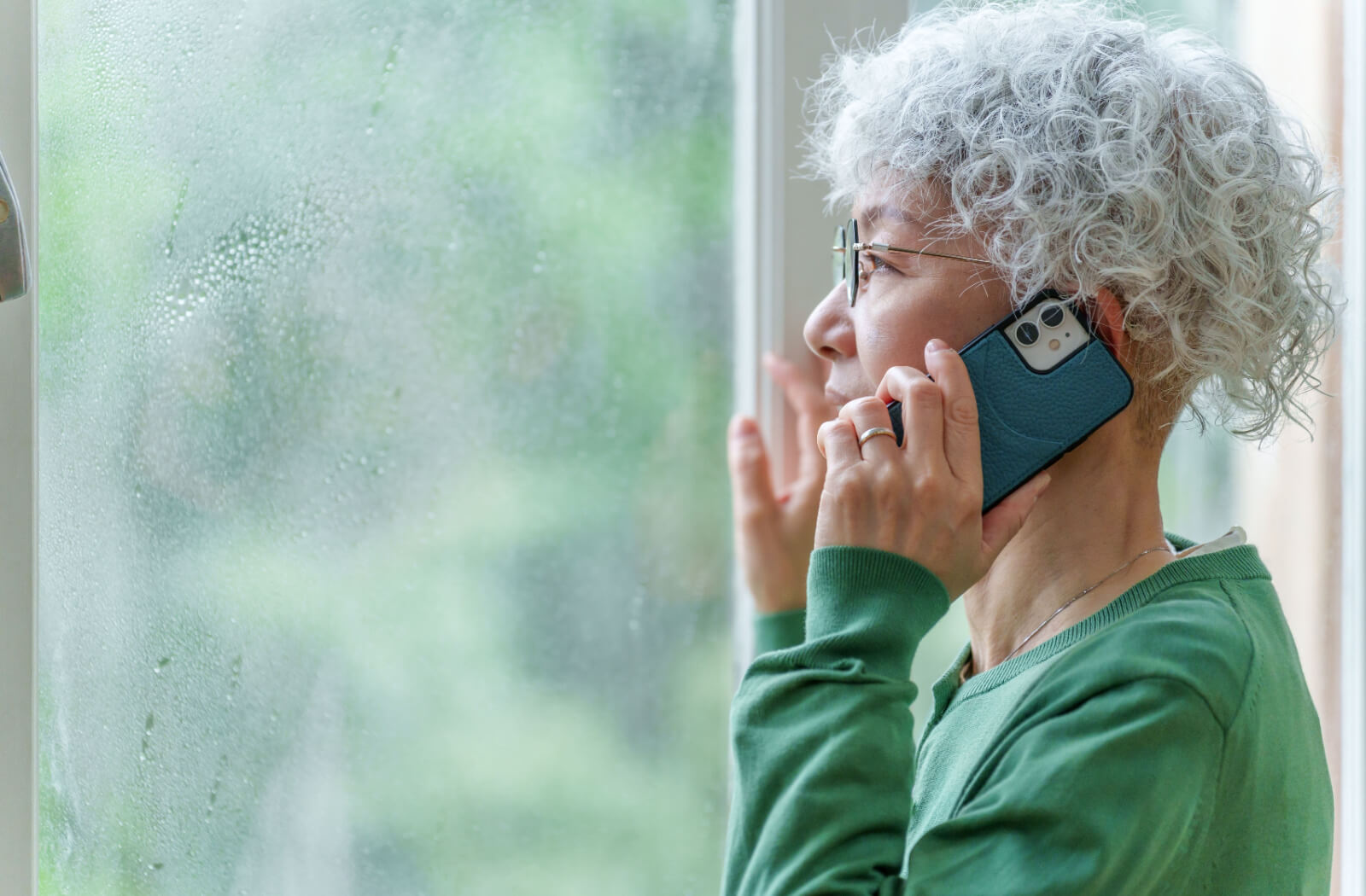 An older adult on the phone with their loved ones while looking out the window at a hurricane.