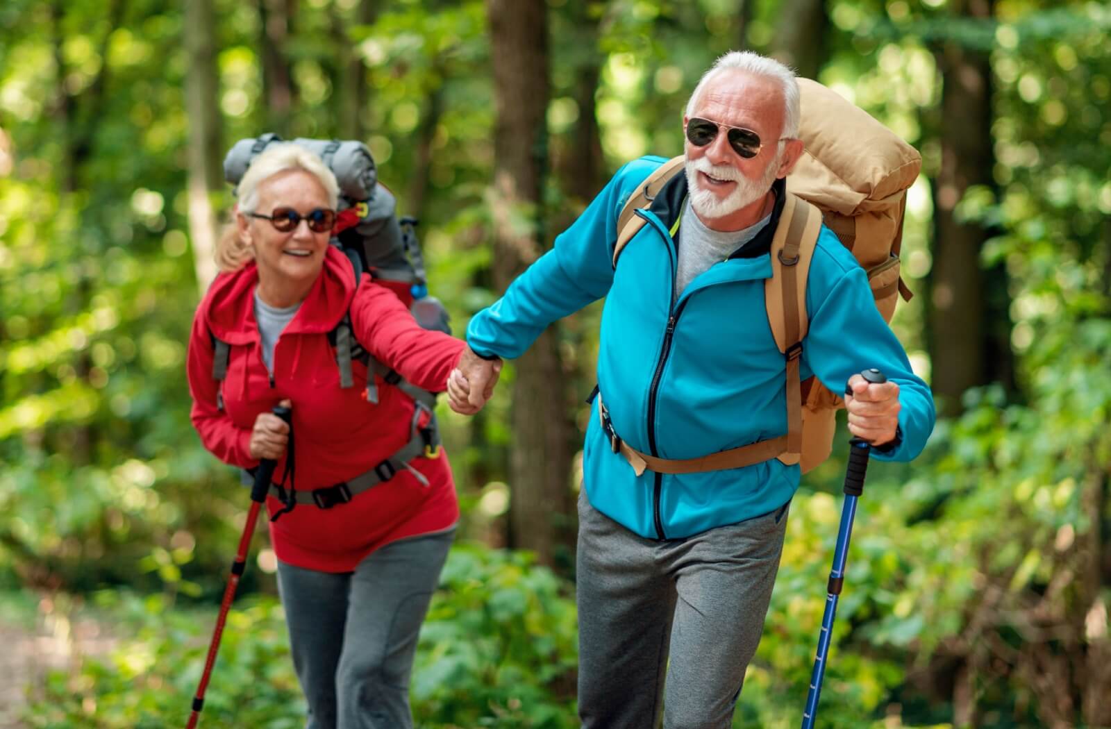A senior couple in vivid jackets with backpacks and walking sticks hold hands while walking through a state park