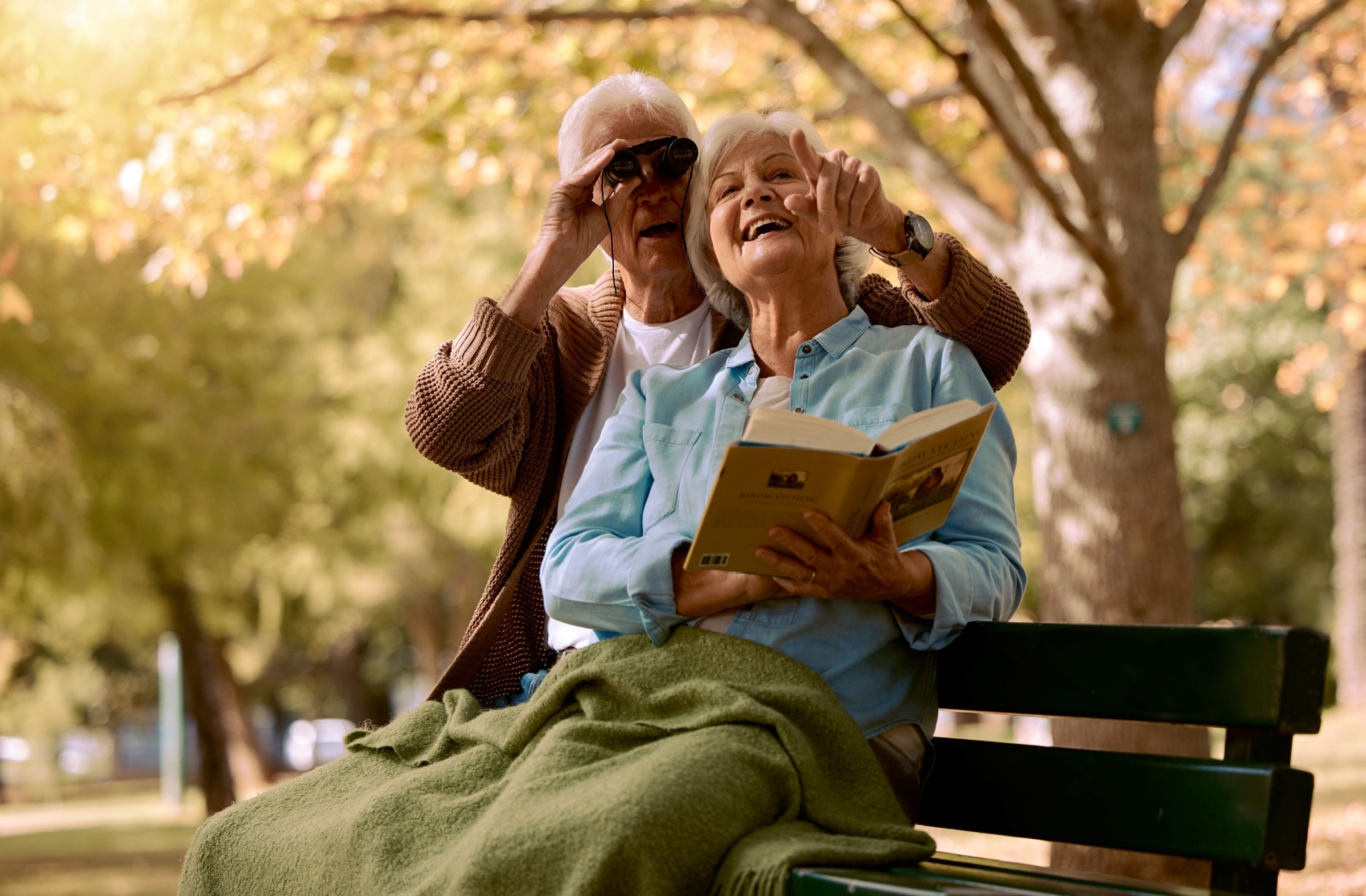 An older couple sits on a bench in a park with a blanket on their lap and a pair of binoculars while looking through a guidebook while birdwatching