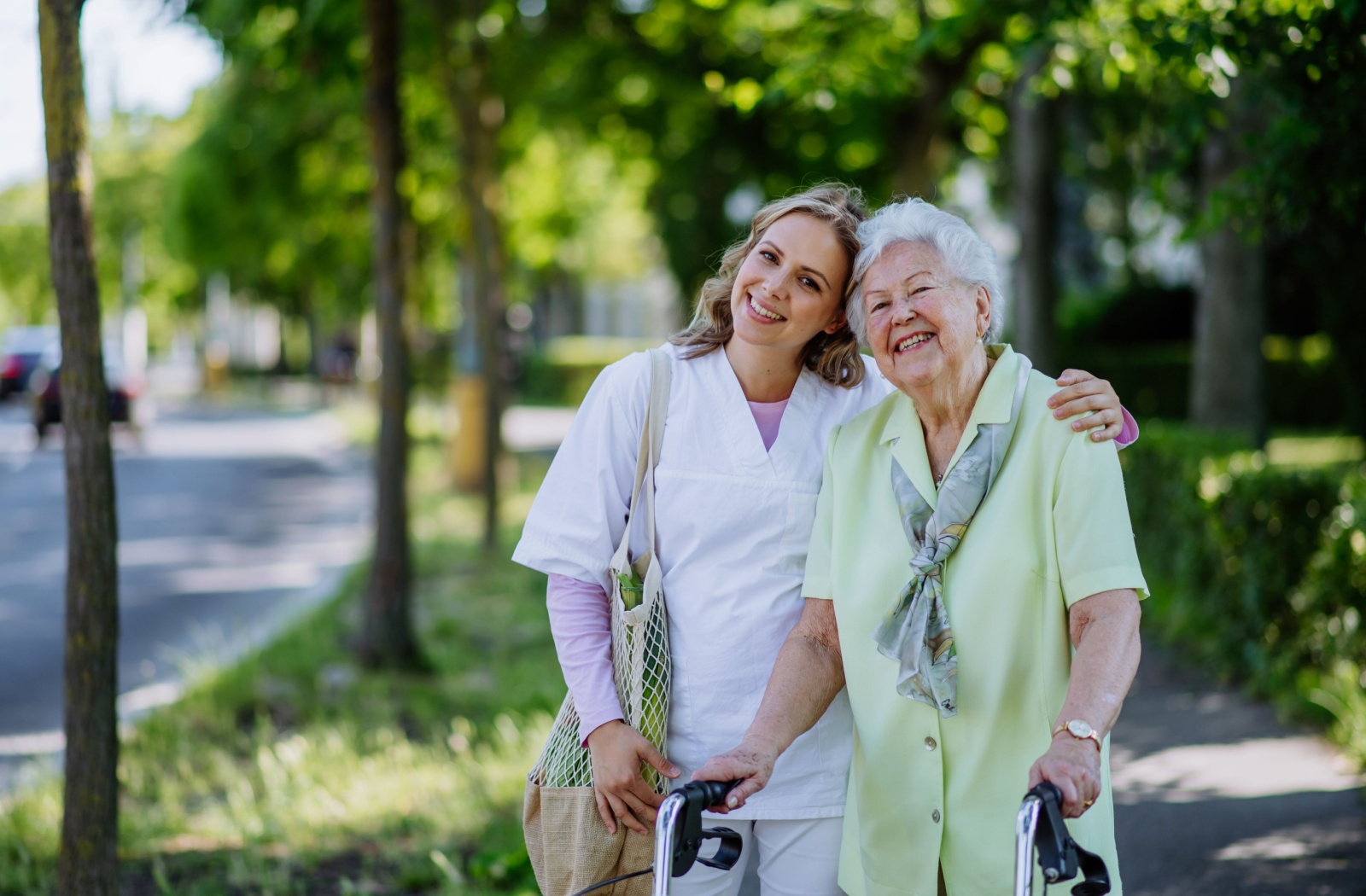 A caregiver puts an arm around an older adult while walking along a nature path outside a senior living community