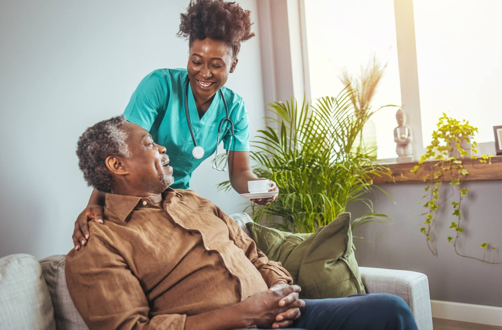 An assisted living caregiver passes a cup of coffee to an older resident sitting on a gray couch.
