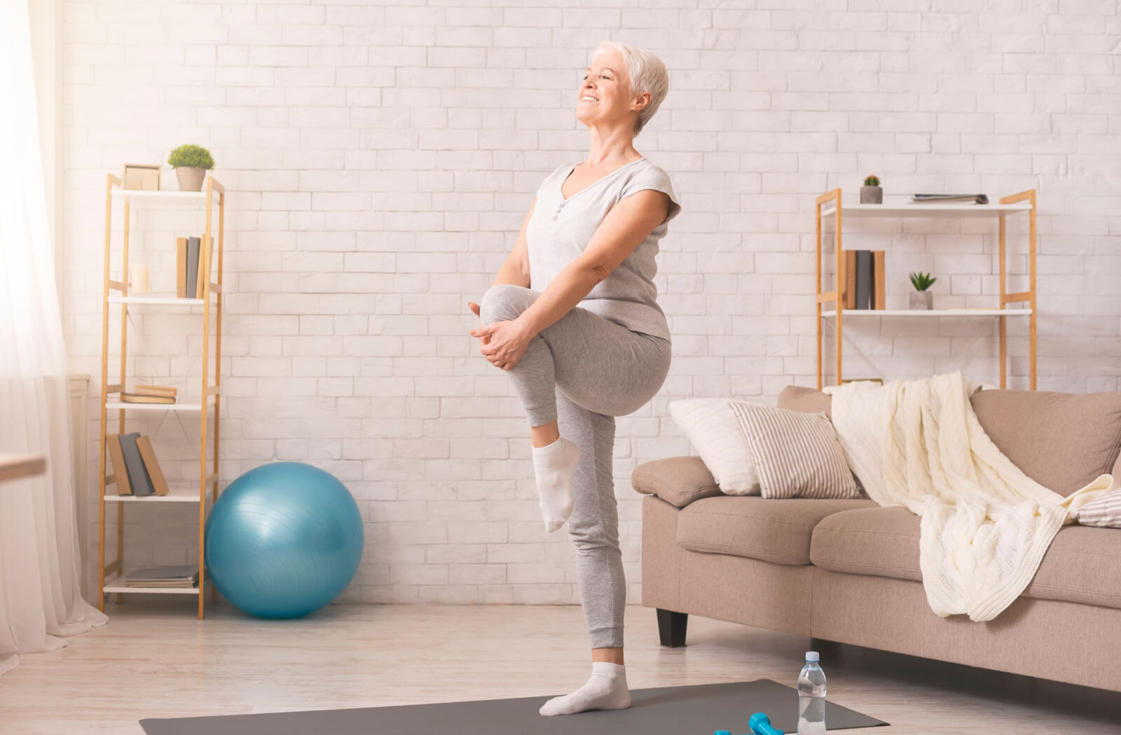 An older adult in a sunlit room holding their knee up and balancing during a leg strengthening exercise.