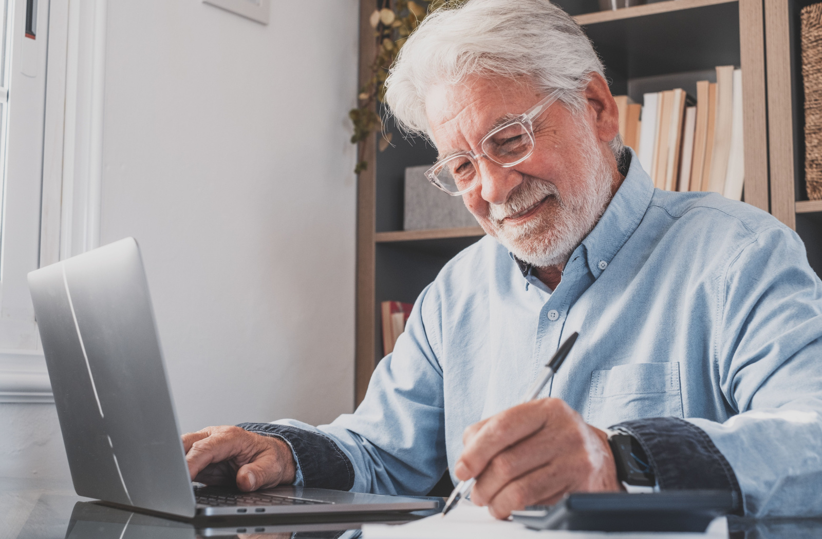 An older adult smiling while using a laptop and notepad to play an online trivia game.