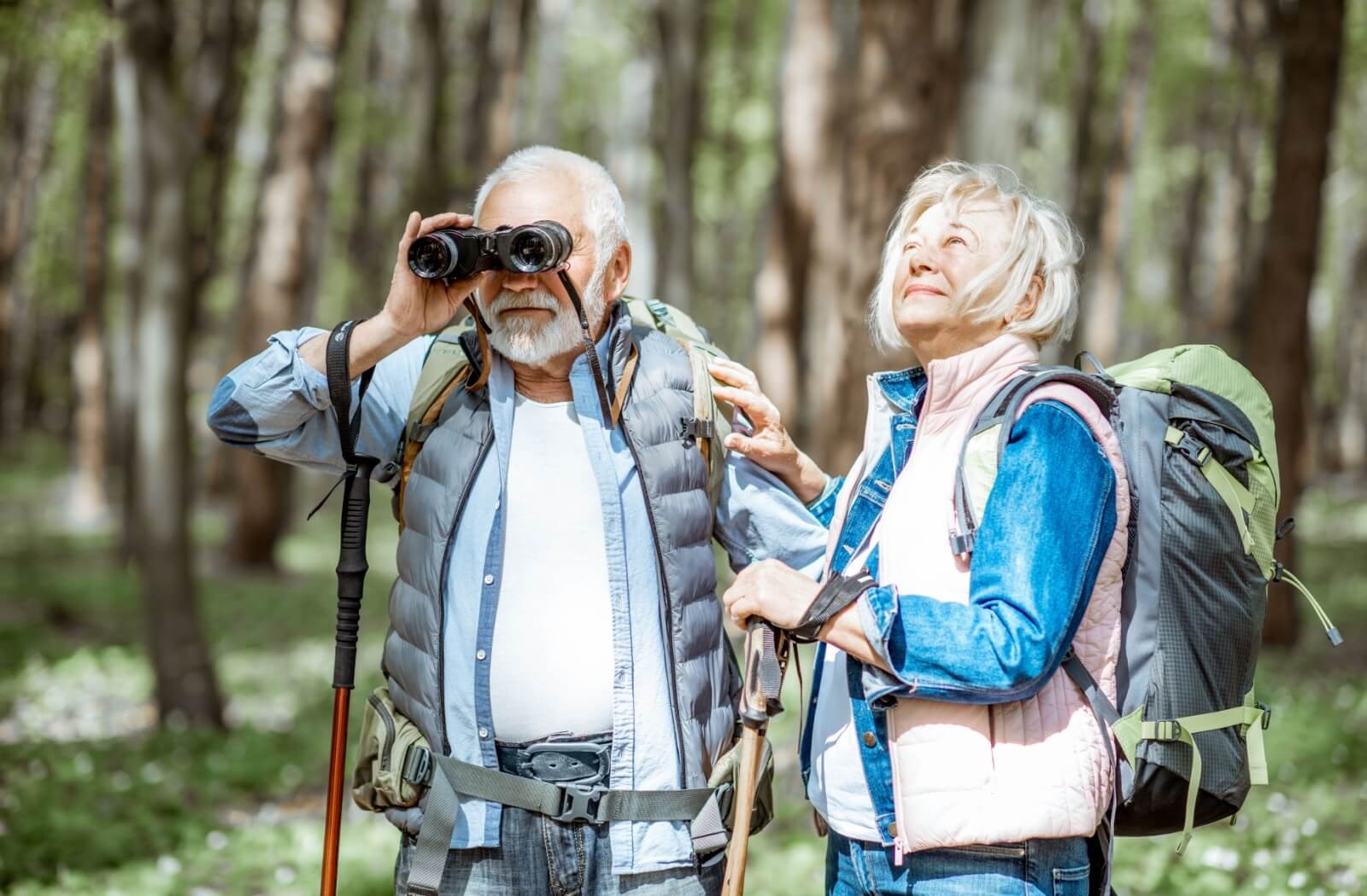 An older adult looks up at the sky while their spouse raises a pair of binoculars while birdwatching in the woods