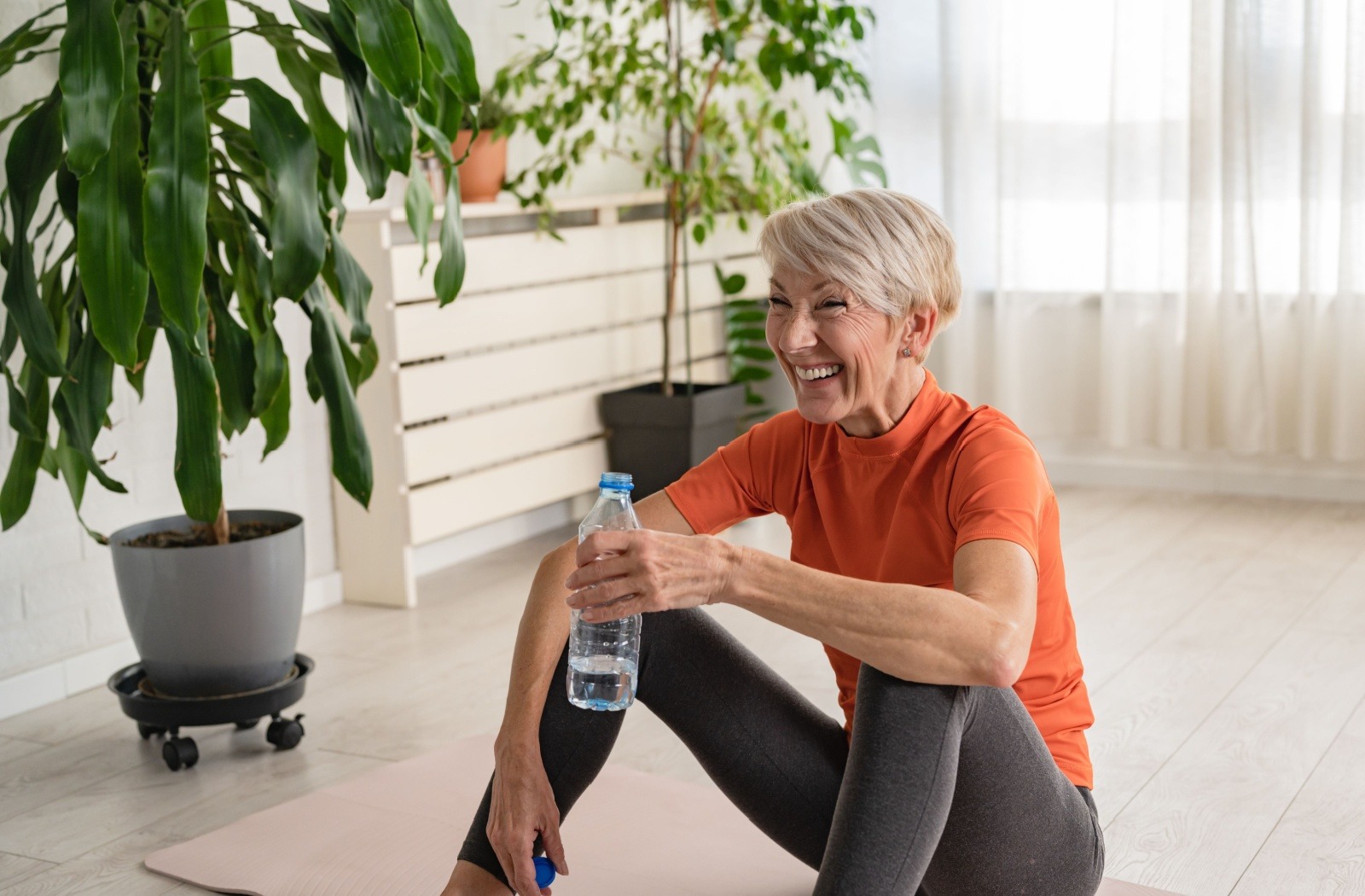 A senior takes a break from a fitness class and drinks a bottle of water while sitting on a yoga mat.
